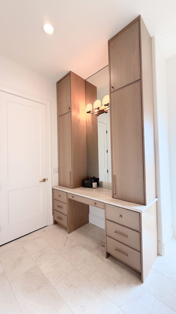 Light oak makeup vanity with tall flanking towers, quartz top, mirror and brass bar pulls in a marble-tiled bathroom