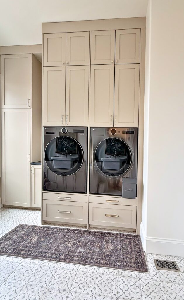 Greige shaker laundry cabinets surrounding a raised washer and dryer with drawers below and patterned tile floor