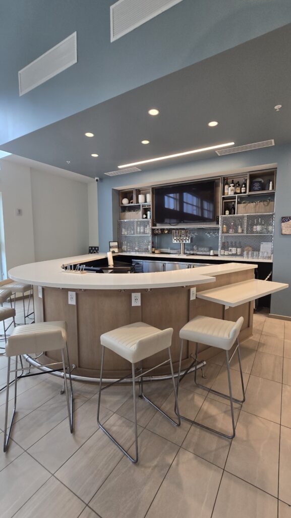 Curved hotel lobby bar with light wood cabinetry, white quartz counter, upholstered stools and backbar shelving