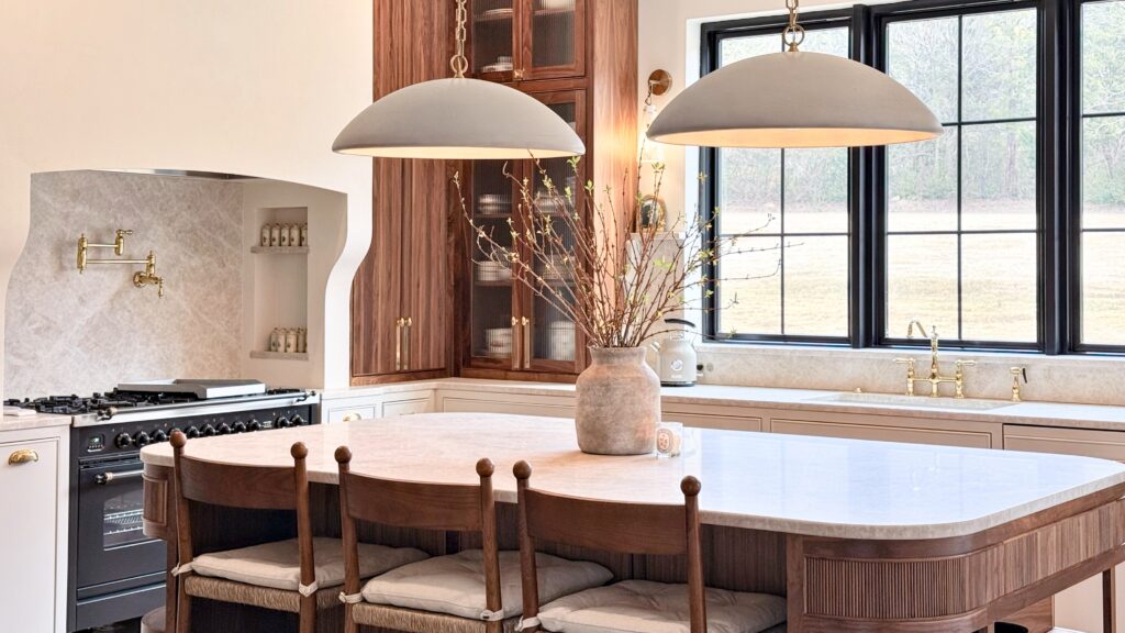 Cream kitchen with fluted walnut island, stone countertop, glass-front wood cabinets, brass fixtures and dome pendants